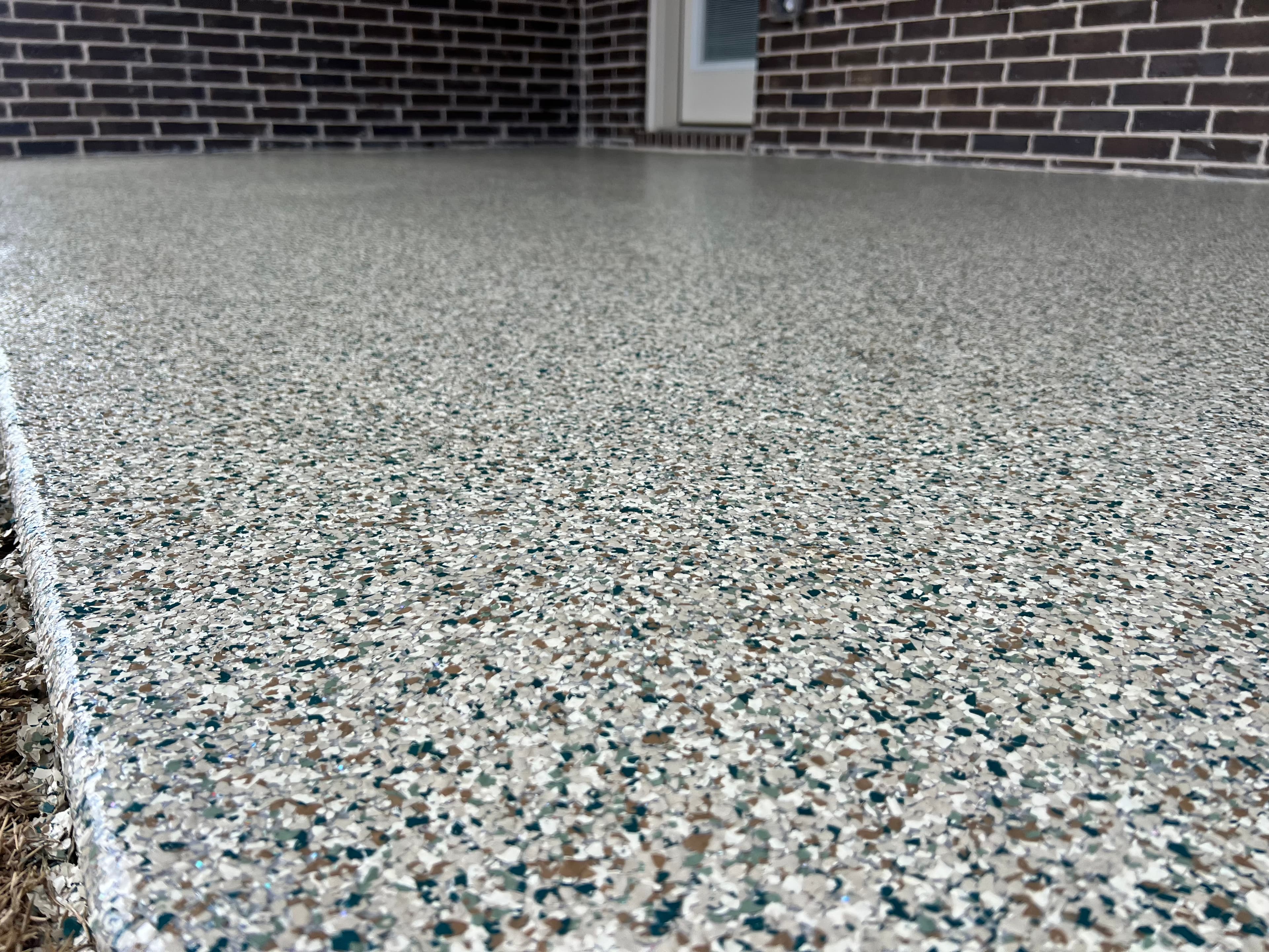 Glossy speckled epoxy floor coating with gray, white, and tan flakes against a brick wall.